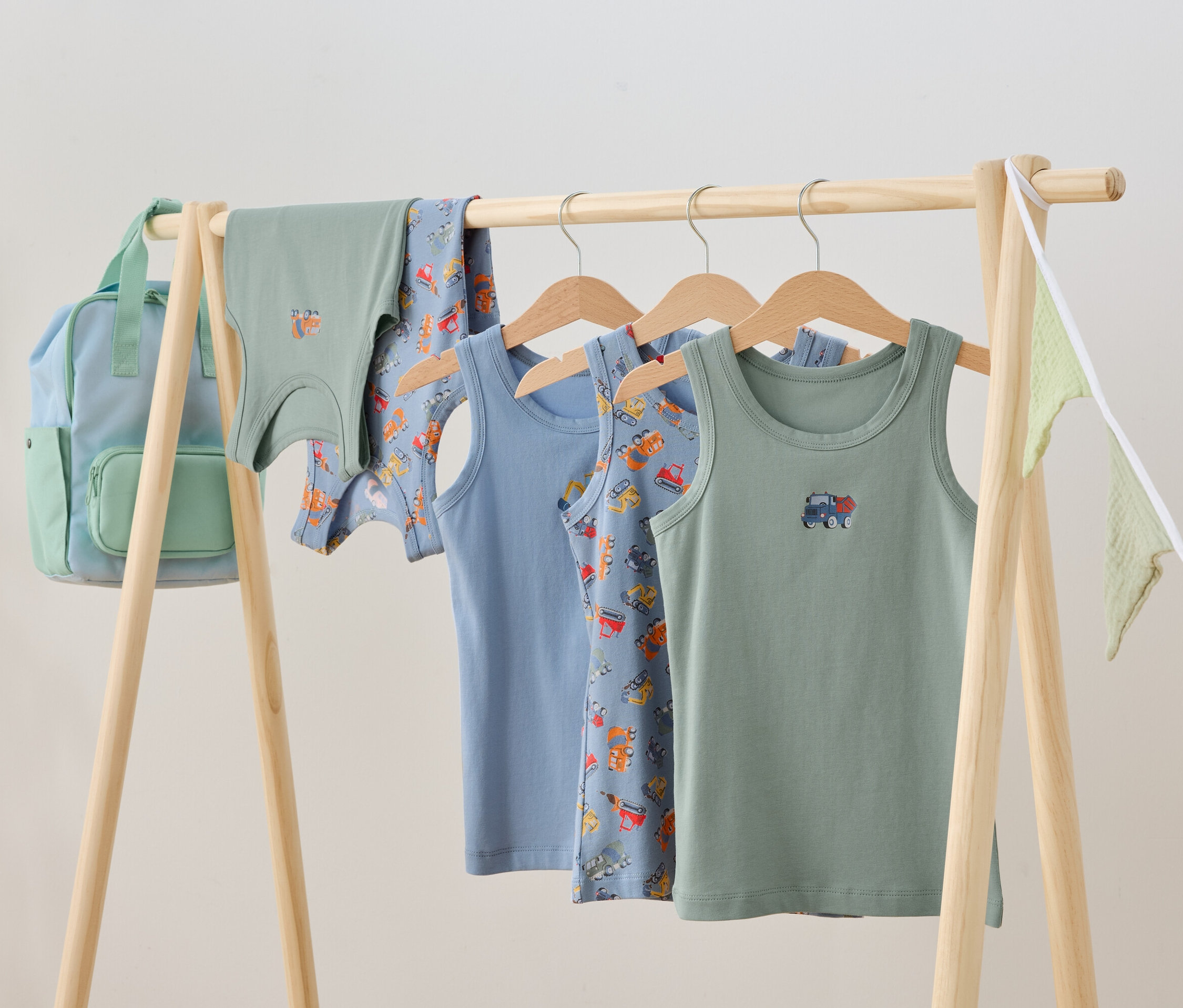 Dětské oblečení visí na dřevěném stojanu, včetně tílek a batohu.