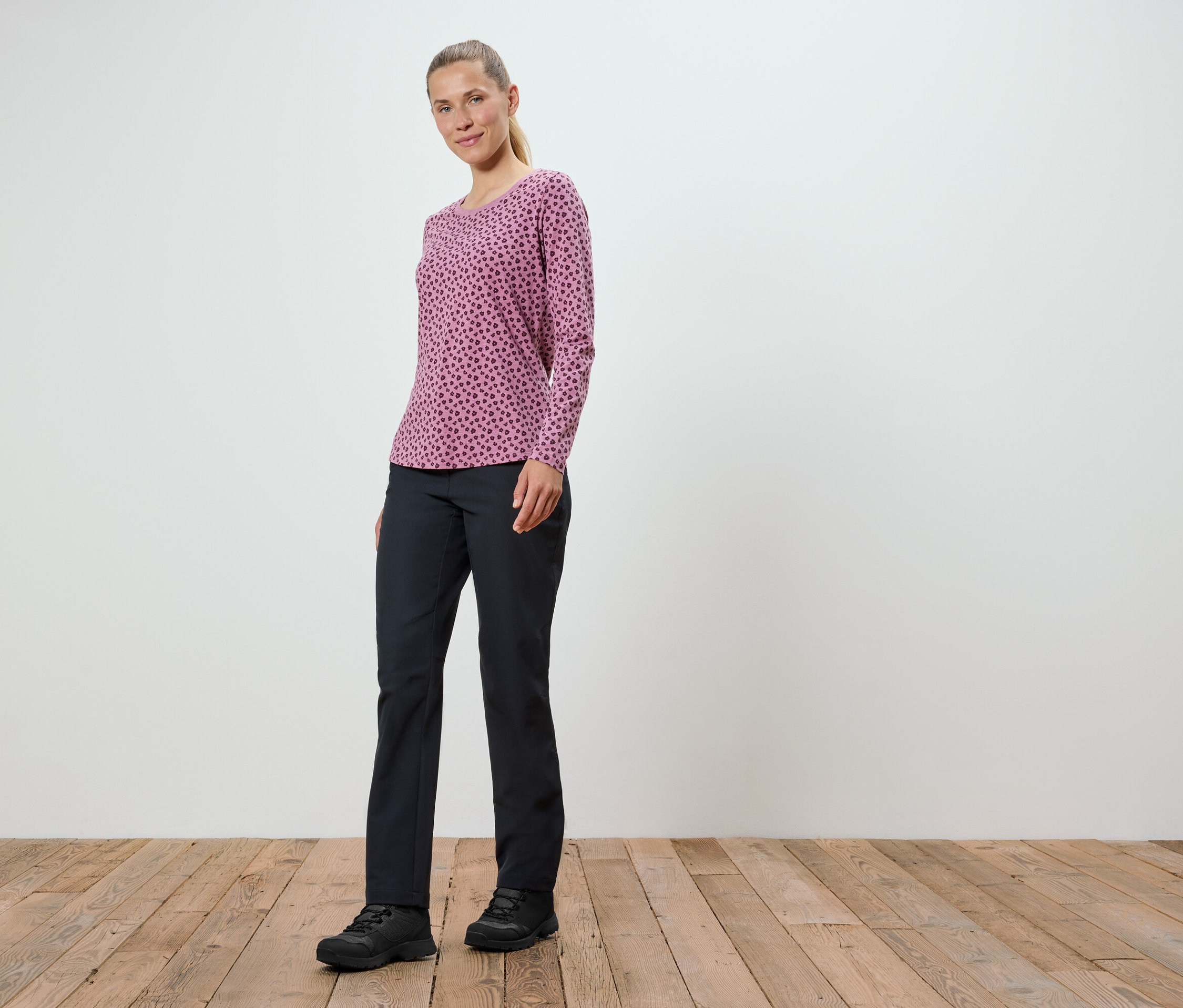 Žena v růžovém tričku s dlouhým rukávem a černých kalhotách stojí na dřevěné podlaze.