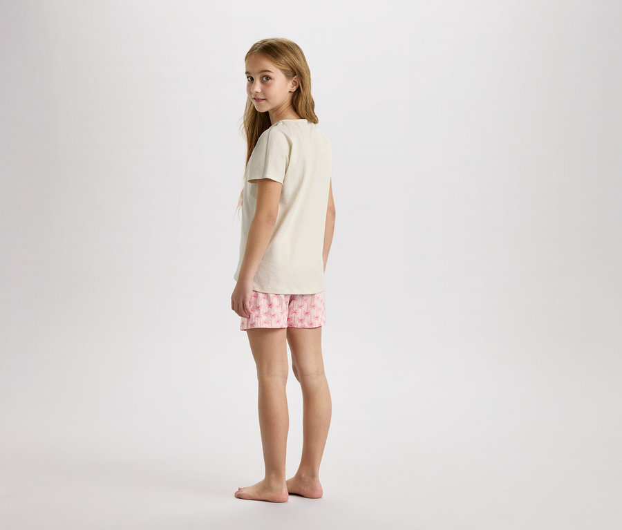 Mladá dívka stojí bosá na neutrálním pozadí v béžovém tričku a růžových šortkách s květinovým potiskem.