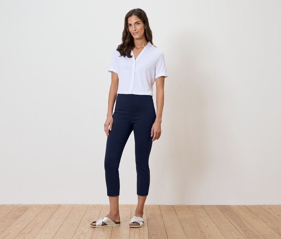 Žena stojí před zdí a má na sobě bílou košili, modré kalhoty a zlaté sandály.