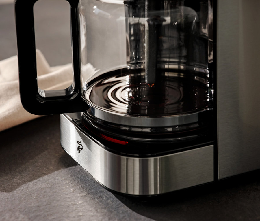 Detailní pohled na kávovar s konvicí na kávu a béžovým hadříkem v pozadí.