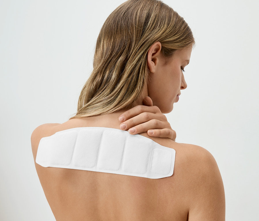 Žena s blond vlasy má na zádech bílou hřejivou náplast a dotýká se krku.