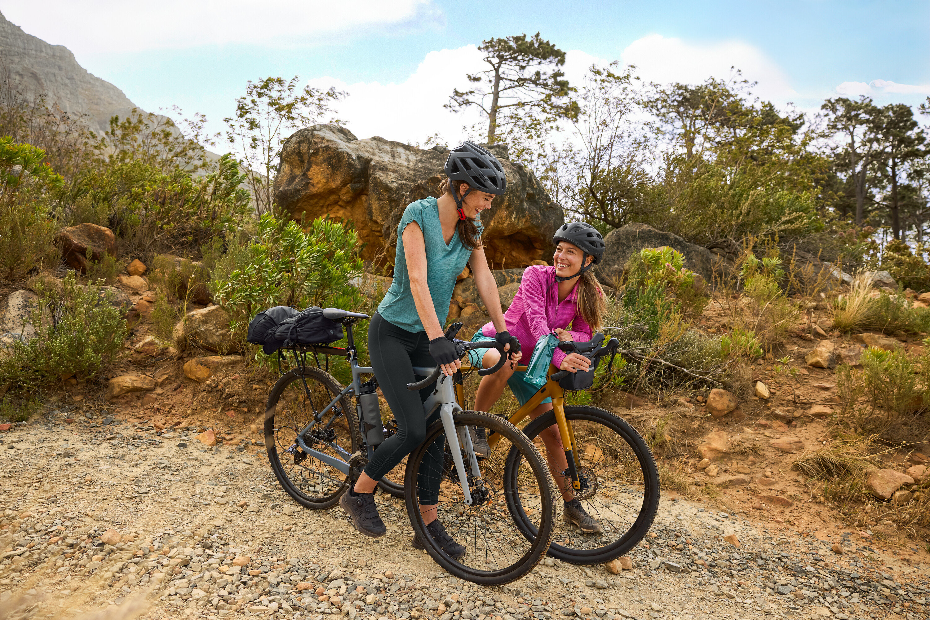 Dvě ženy s helmami stojí s koly na štěrkové cestě v kopcovitém terénu. Na kole je připevněna láhev na pití.