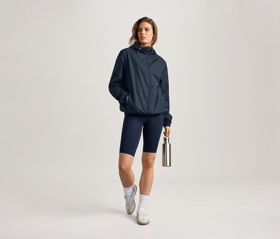 Žena v bundě do deště, cyklistických šortkách a s lahví na vodu. Má na sobě ponožky a tenisky.