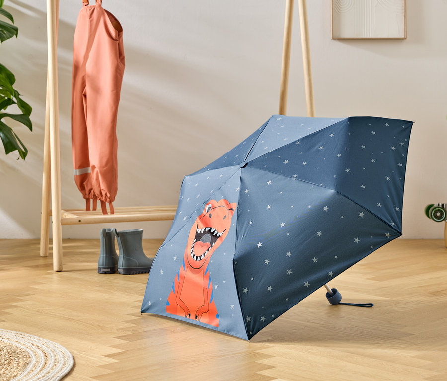 Modrý deštník s motivem dinosaura stojí na dřevěné podlaze, vedle něj je věšák s oblečením do deště a gumáky.