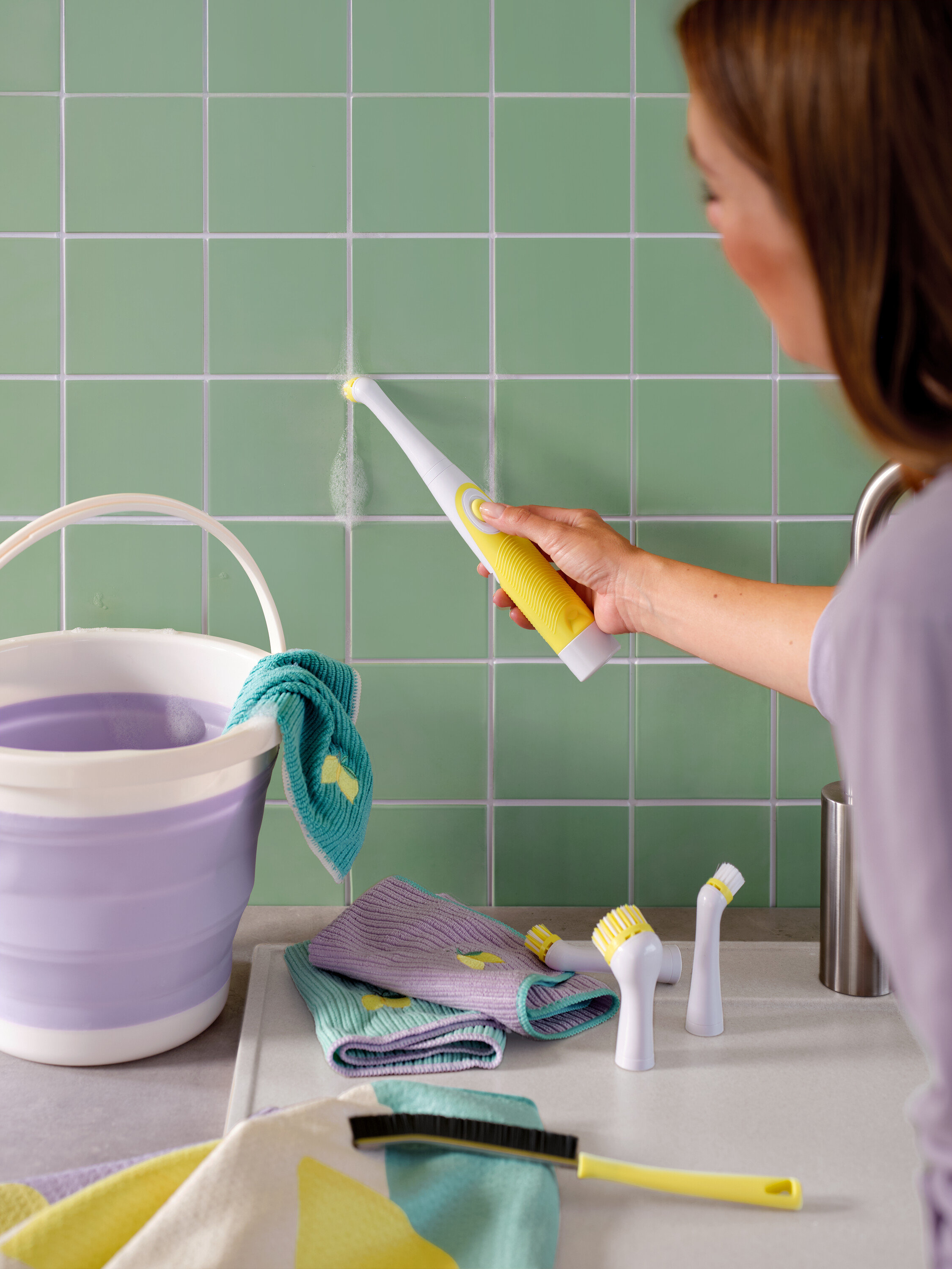 Žena čistí zelené dlaždice žluto-bílým kartáčem, viditelné je také vědro a hadry.