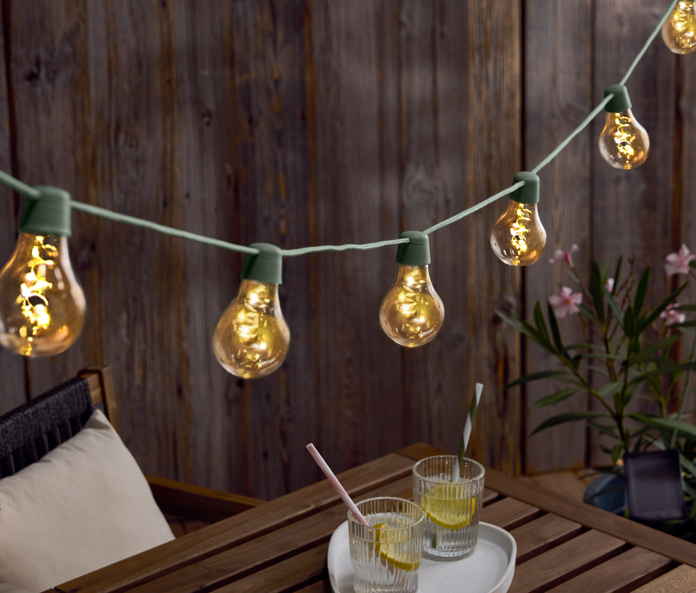 Světelný řetěz se žárovkami visí nad dřevěným stolem s nápoji a kloboukem.