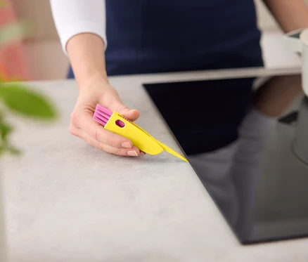 Žena čistí černou varnou desku žluto-růžovým čisticím nástrojem.