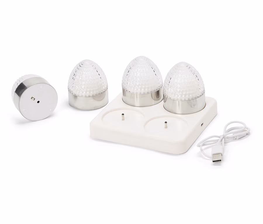 Čtyři malé kulaté lampy se stříbrnými základnami a fasetovanými, průhlednými skleněnými kopulemi stojí na bílé nabíjecí stanici s USB kabelem.