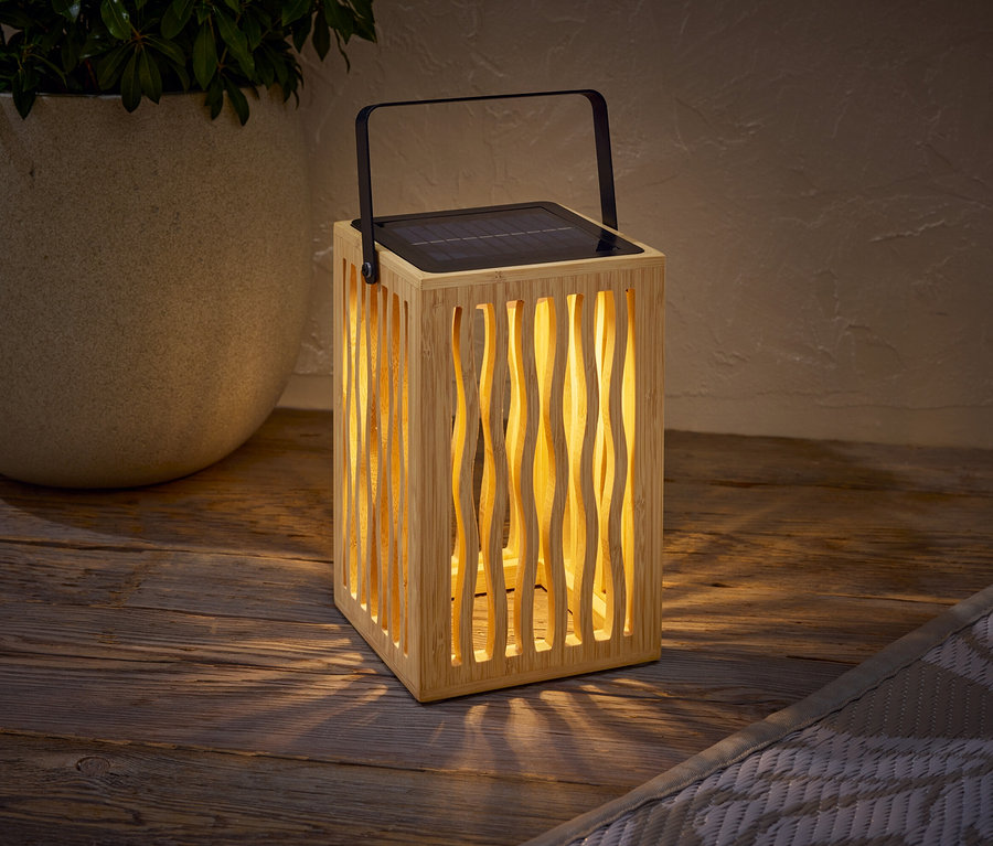 Dřevěná solární lampa s černou rukojetí stojí na dřevěném stole vedle květináče s rostlinou.