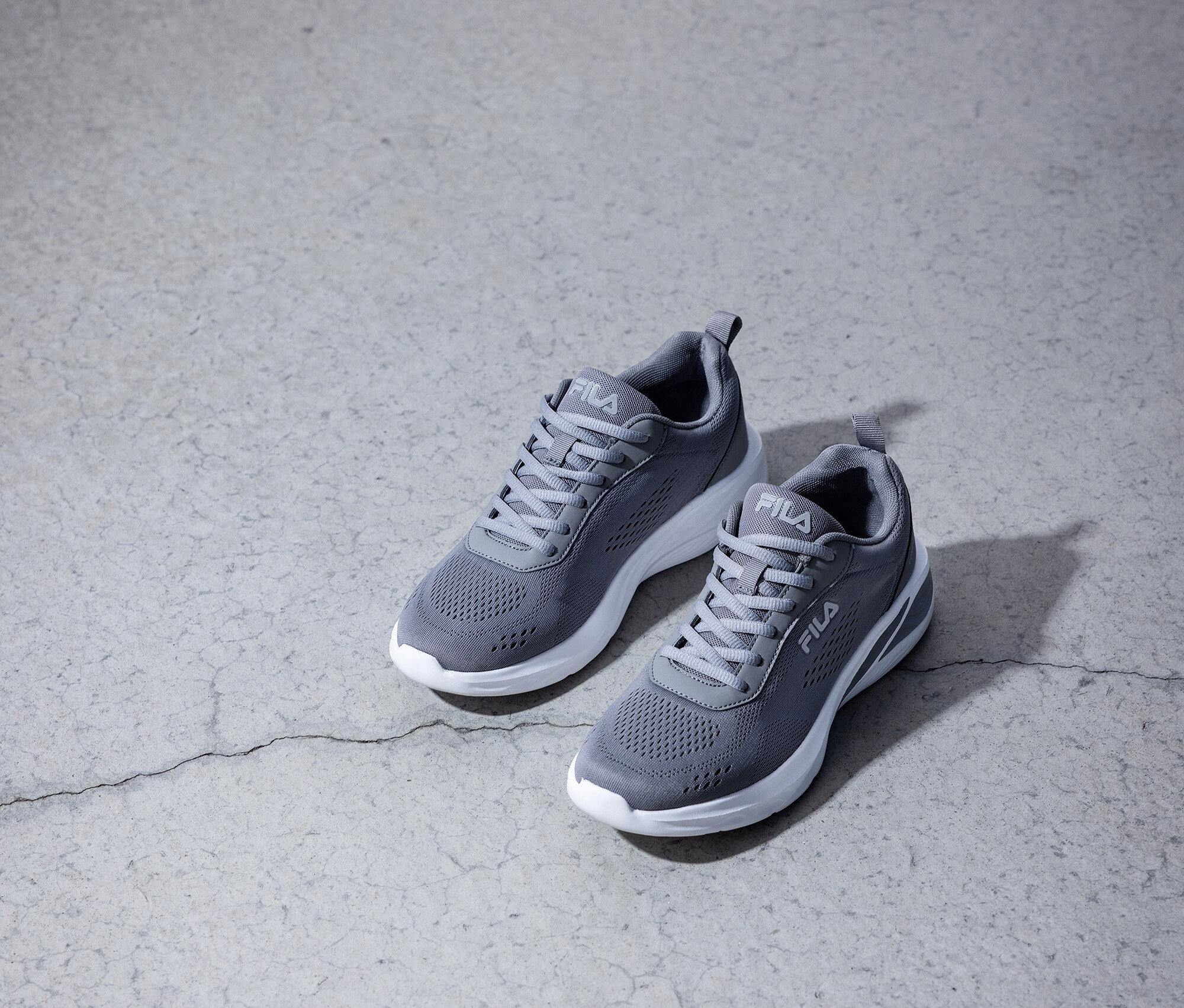 Dva šedé tenisky Fila s bílými podrážkami stojí na šedé betonové podlaze.
