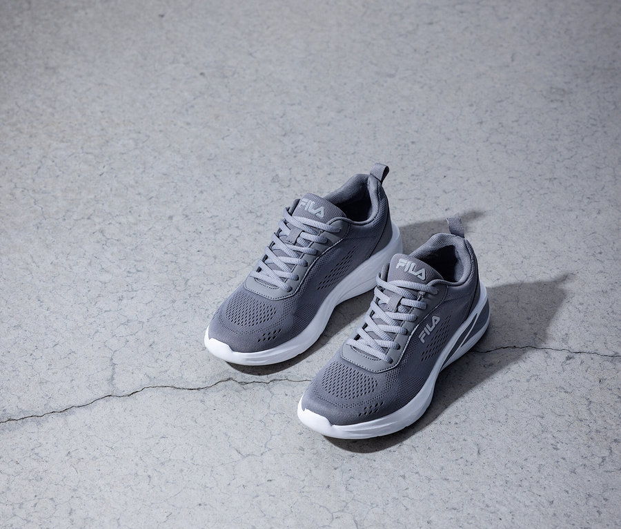 Dva šedé tenisky Fila s bílými podrážkami stojí na šedé betonové podlaze.