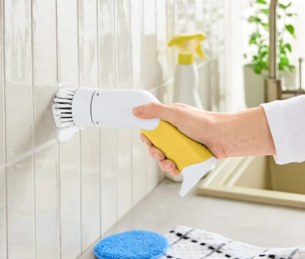 Ruka drží elektrický čisticí kartáč se žlutou rukojetí a bílou hlavicí, čistící dlaždice.