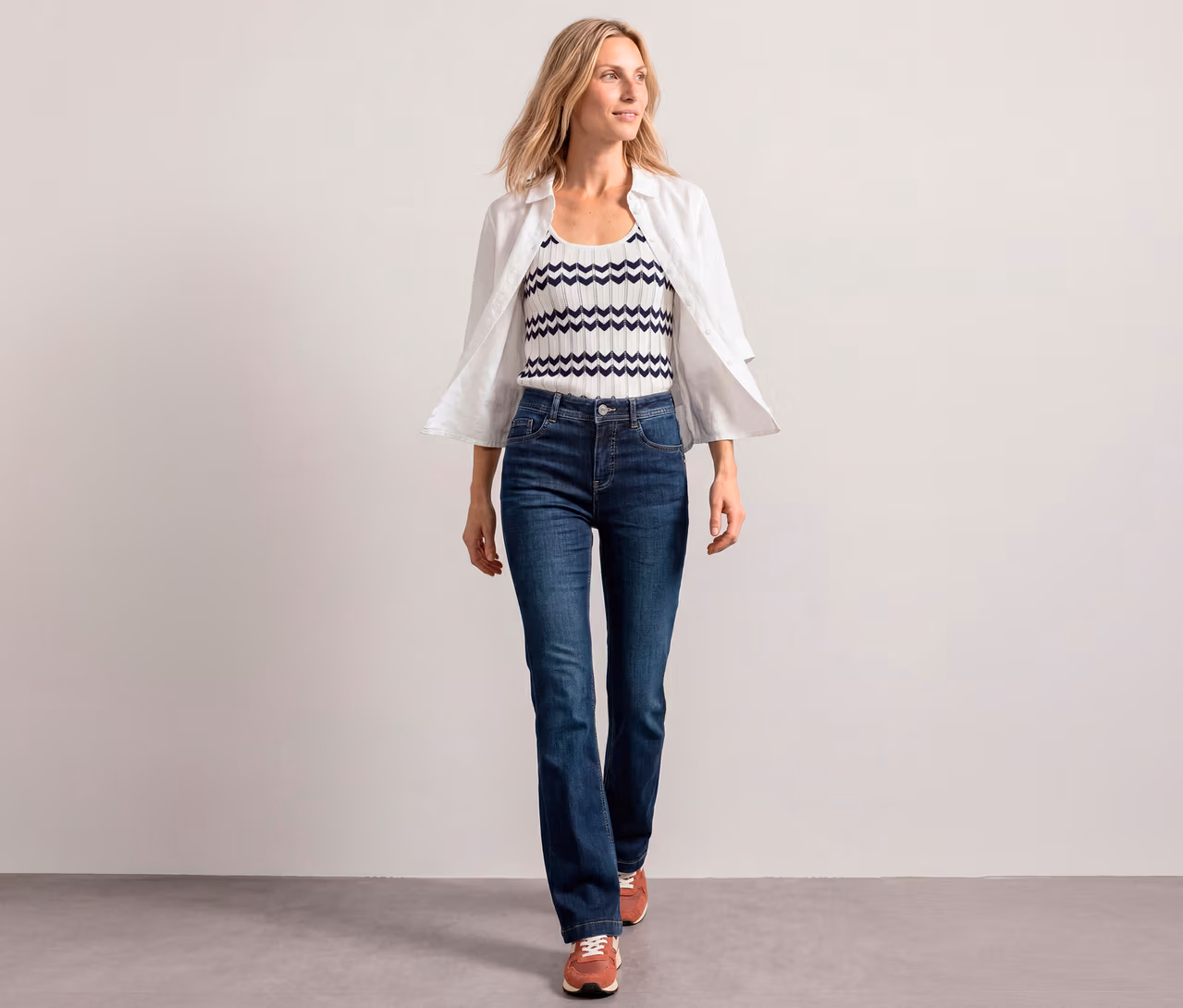 Žena kráčí vpřed, má na sobě bílou rozepnutou košili, bílý a tmavě modrý top se vzorem cikcak a džíny bootcut Street One.