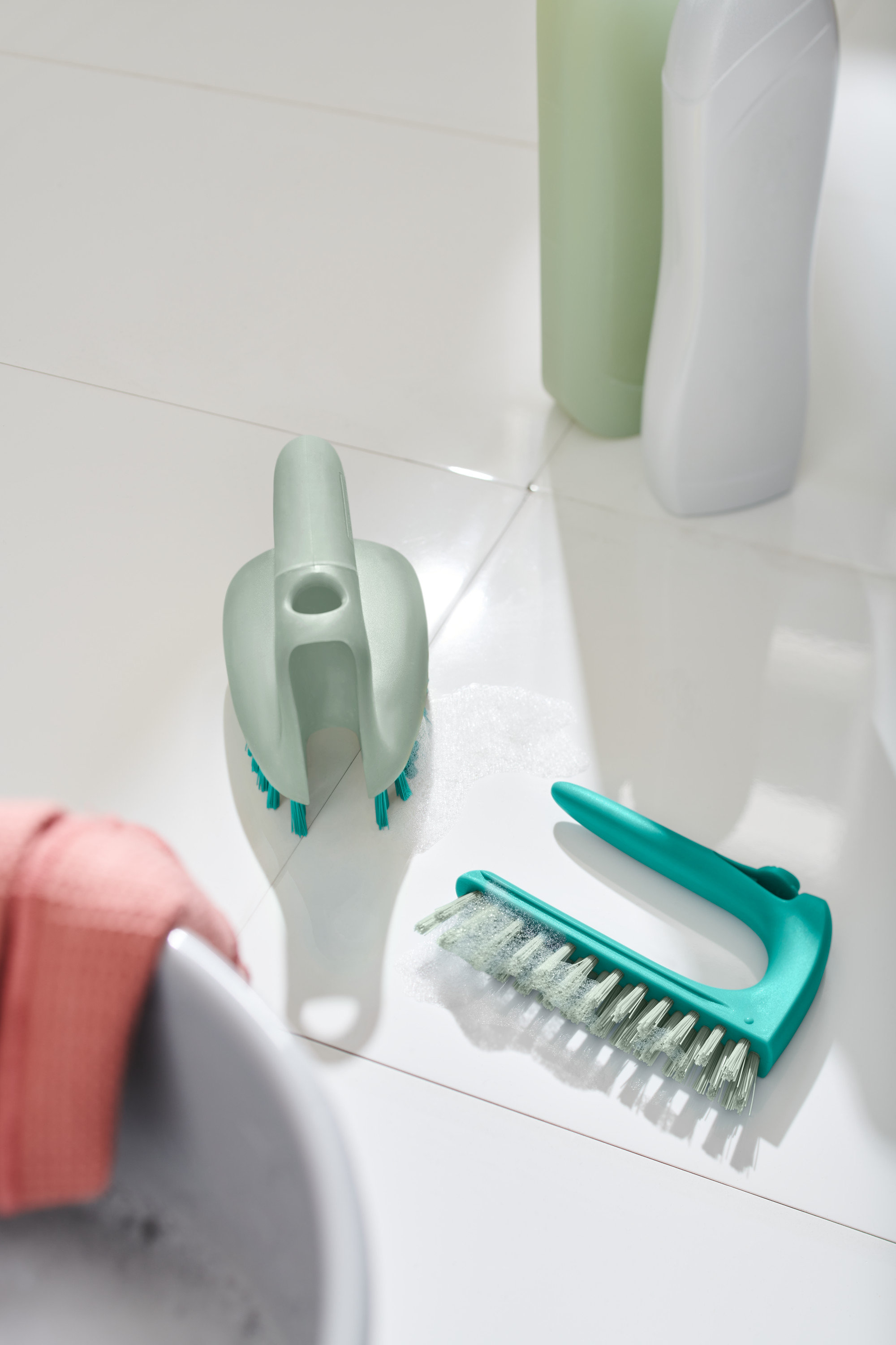 Dva kartáče na podlahu 2 v 1 leží na bílých dlaždicích vedle umyvadla.