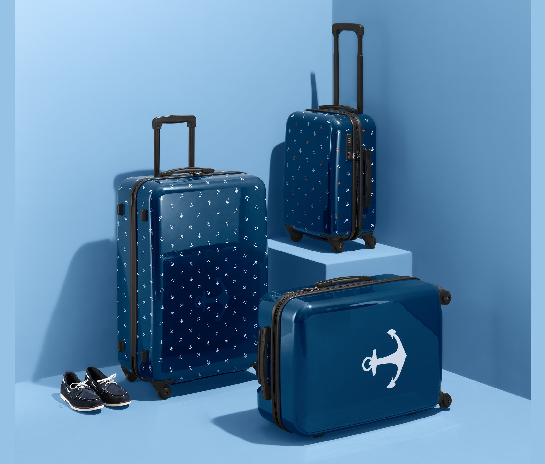 Tři modré kufry z tvrdé skořepiny (velký, střední a malý) s potiskem kotvy a modré boty na pozadí modrého studia.