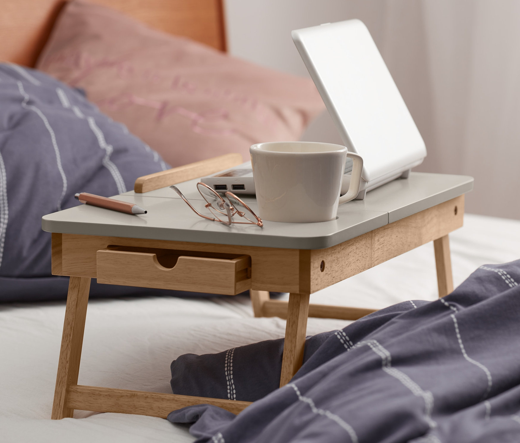 Na posteli je stolek do postele s notebookem, hrnkem, brýlemi a tužkou.