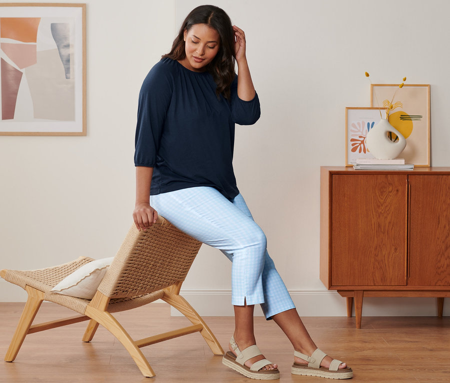 Žena sedí na židli a má na sobě tmavě modré tričko s 3/4 rukávem, 7/8 strečové kalhoty světle modro-bílé a kožené sandály na platformě.