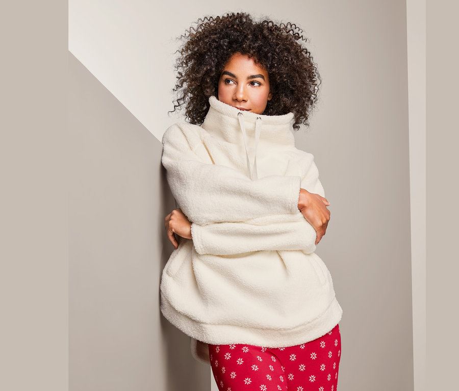 Žena s kudrnatými vlasy stojí opřená o zeď a má na sobě hebkou mikinu a červené kalhoty.