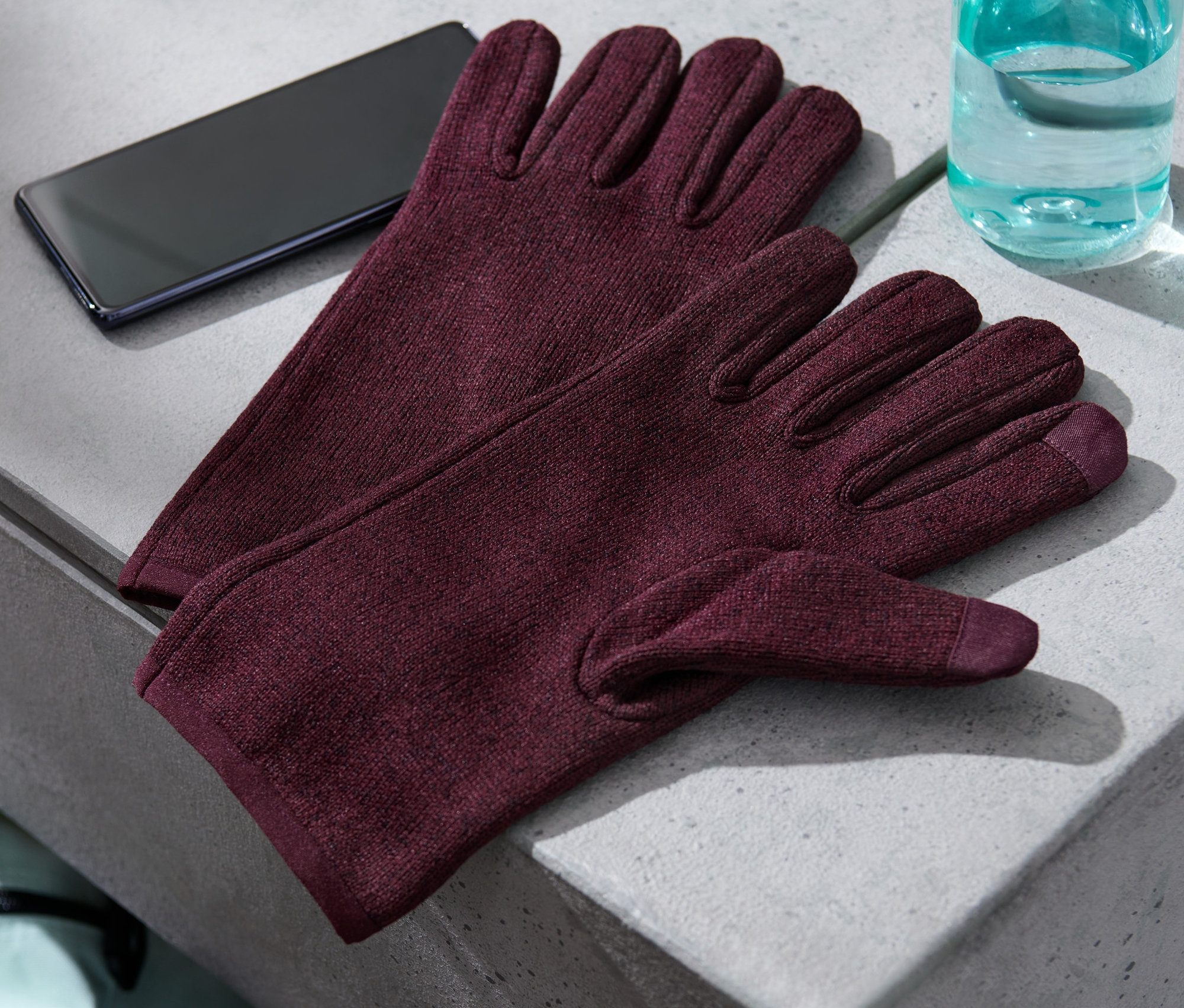 Na šedé ploše leží pár rukavic z pleteného fleecu, mobilní telefon a láhev s vodou.