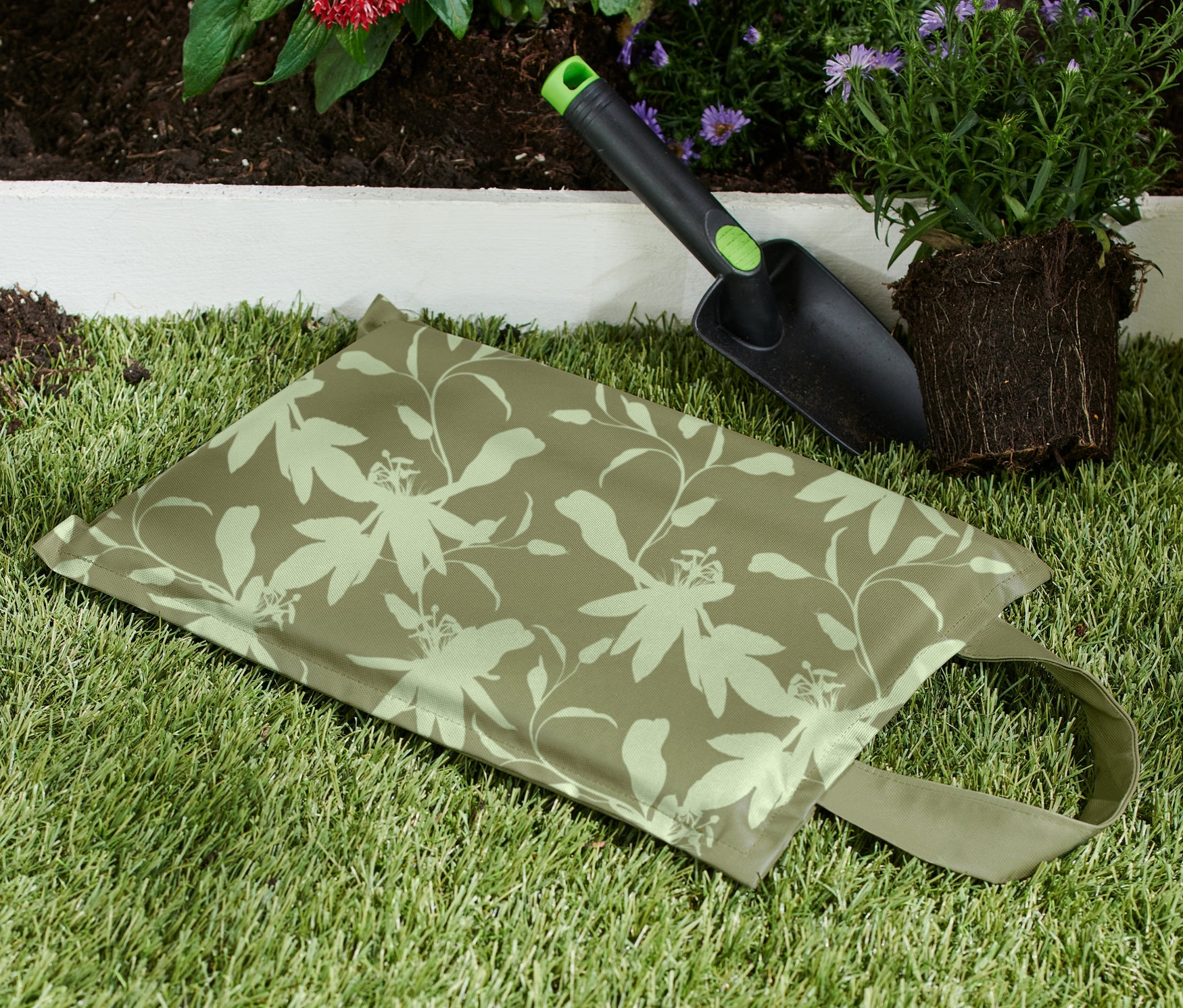 Zahradnický polštářek na klečení leží na trávě vedle lopatky a rostliny.