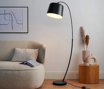 Černá prohnutá stojací lampa z kovu stojí vedle béžové pohovky.