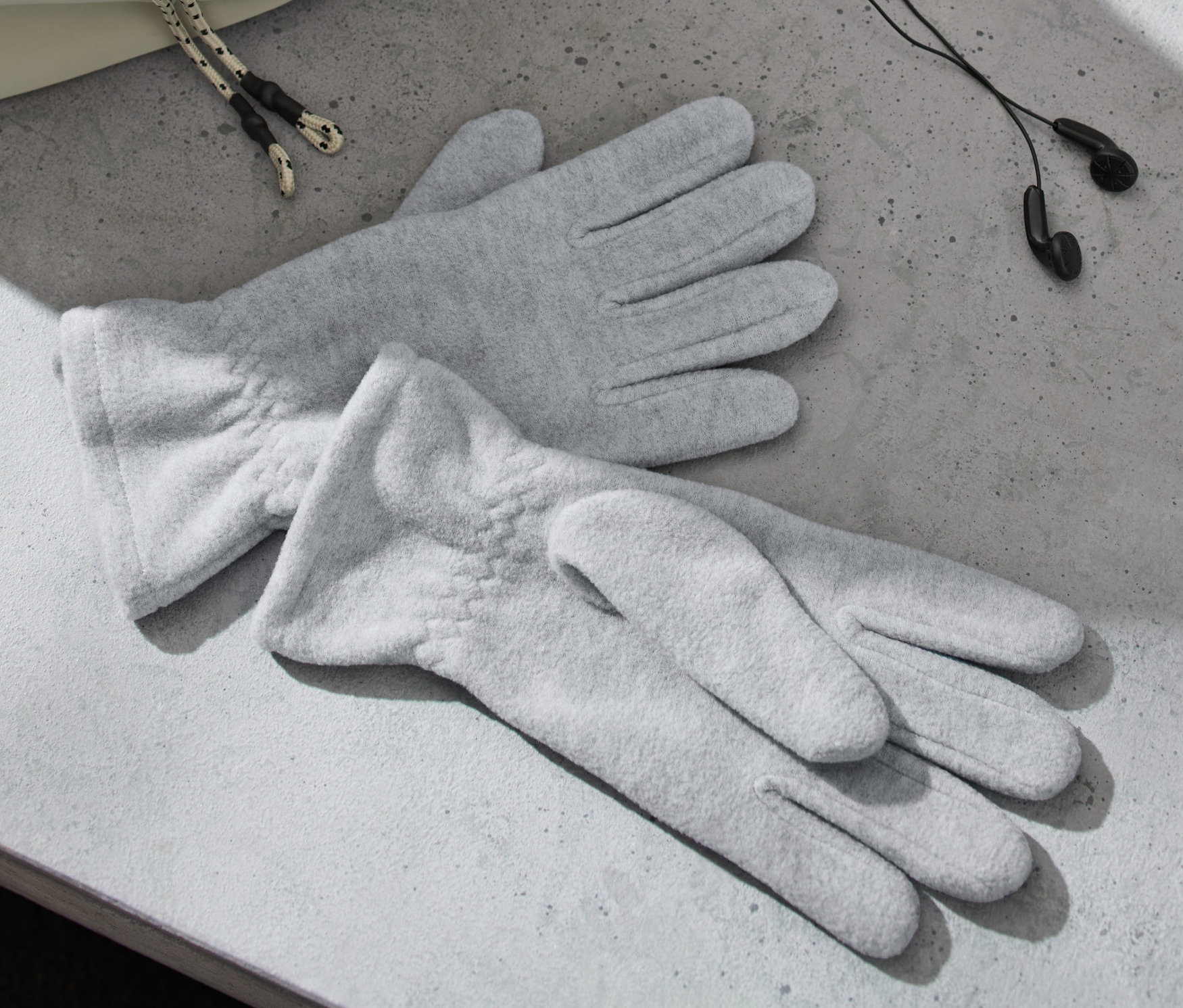 Dvoje šedé fleecové rukavice leží na šedém povrchu vedle sluchátek a tašky.