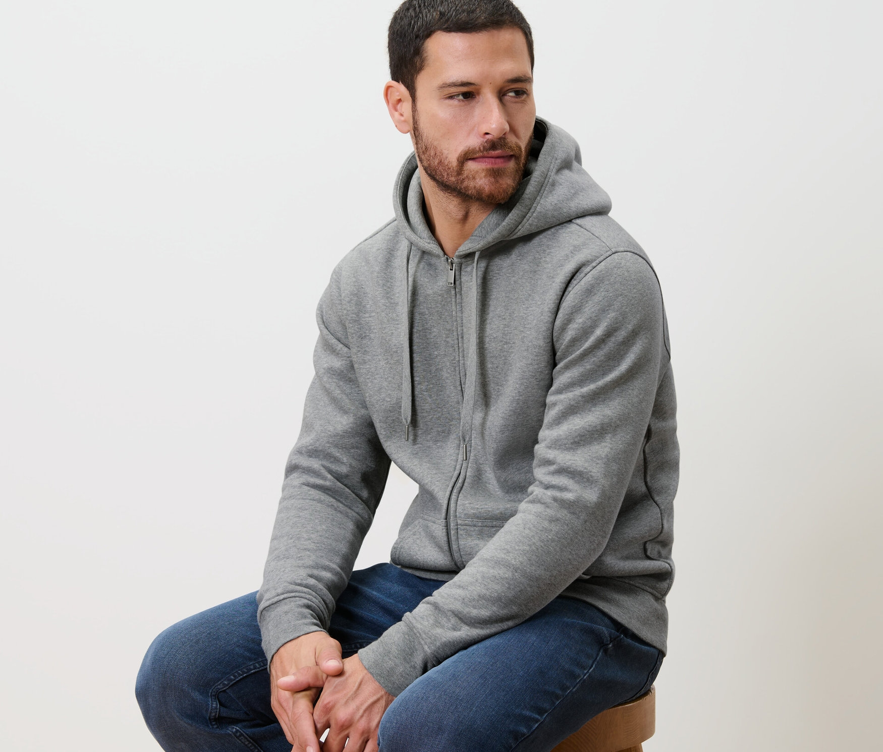 Muž sedí na stoličce a má na sobě šedou bundu s kapucí.