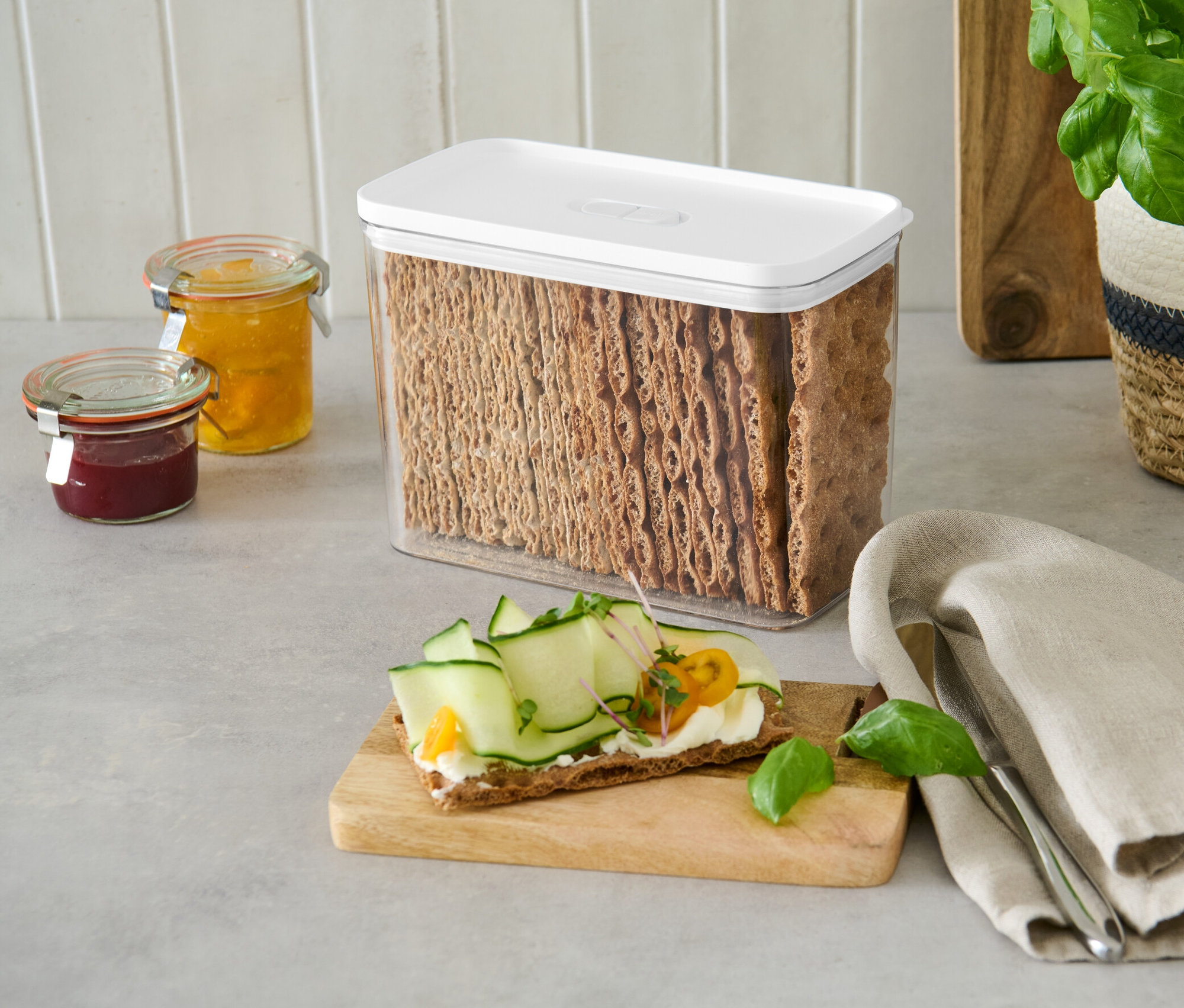 Dóza na uchovávání potravin s bílým víkem plná chlebových krekrů stojí na lince vedle sklenic s džemem.