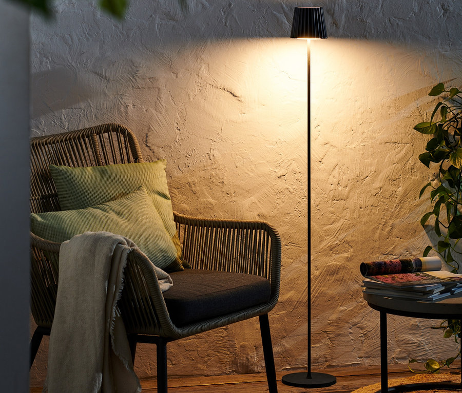 Černá bezdrátová stojací LED lampa stojí vedle proutěného křesla s polštáři.