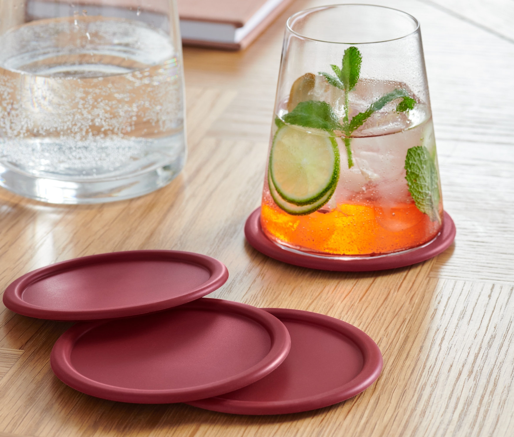 Sklenice s nápojem stojí na jednom ze čtyř bordó silikonových podtácků. Další tři podtácky leží vedle sebe na dřevěném stole. V pozadí je karafa s vodou a notes.