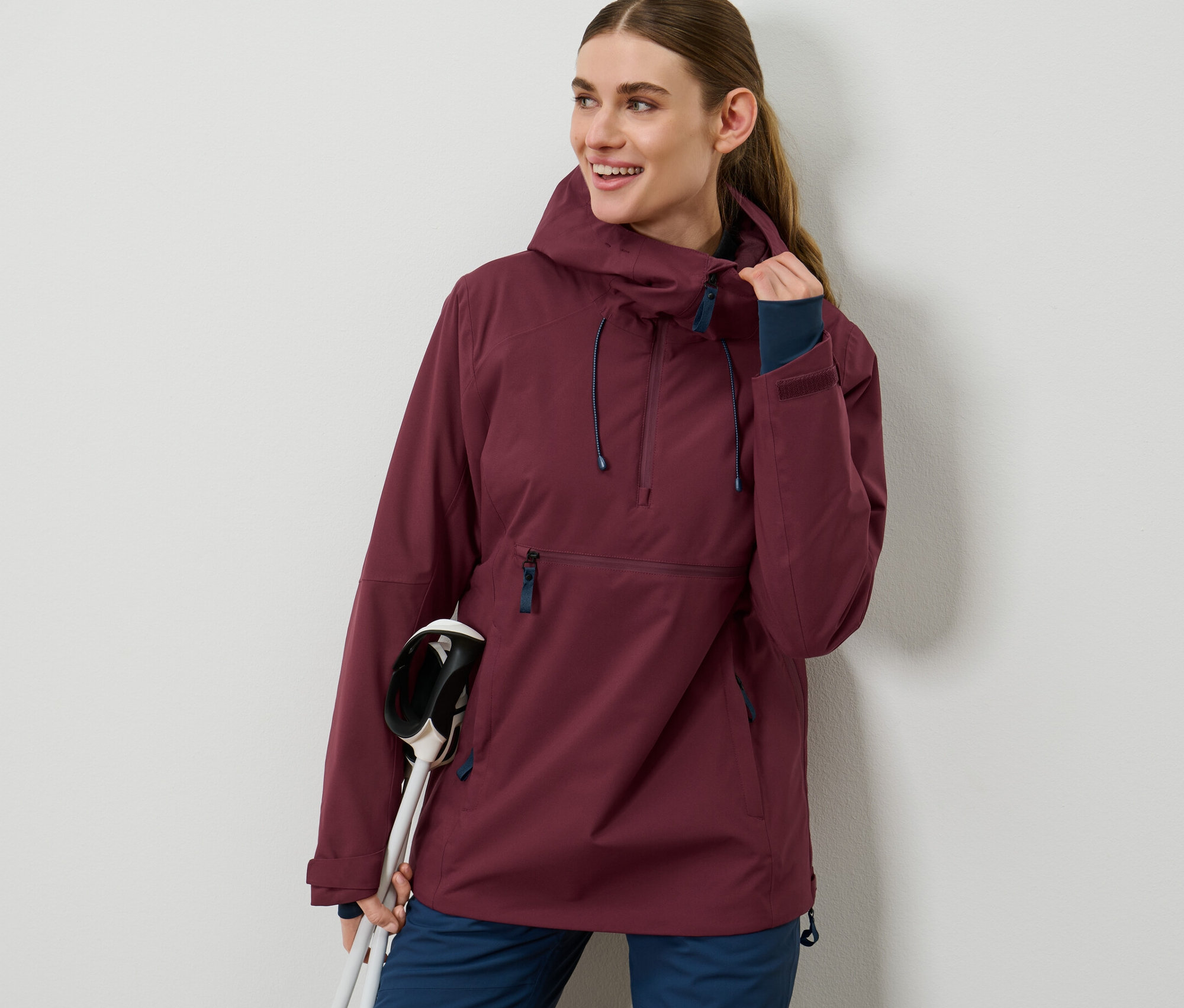 Žena má na sobě bordó lyžařskou bundu s polovičním zipem a drží lyžařské hůlky.