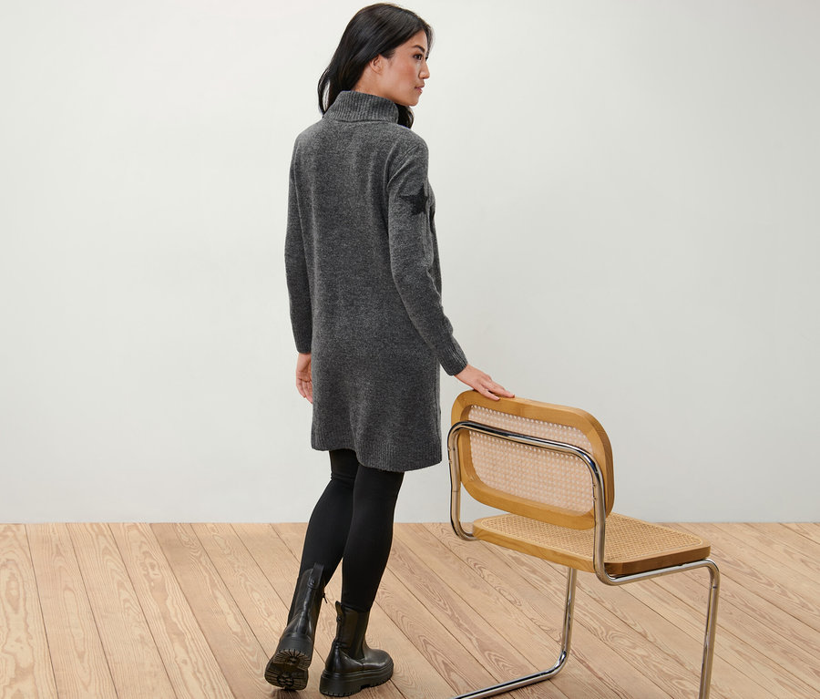 Žena stojí zády a pózuje v pletených šatech s rolákem.