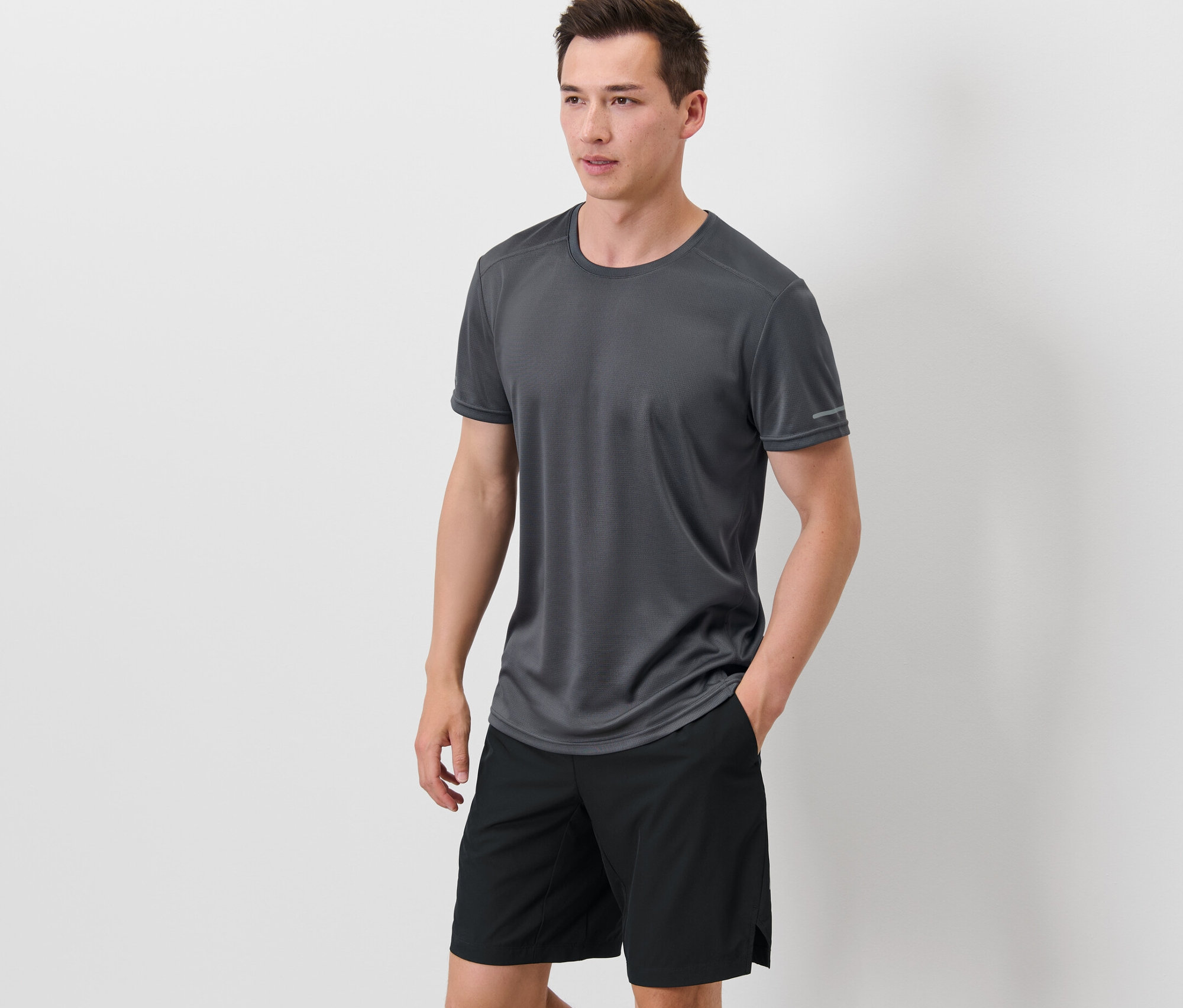 Muž stojí a má ruce v kapsách černých šortek, na sobě má šedé funkční triko.