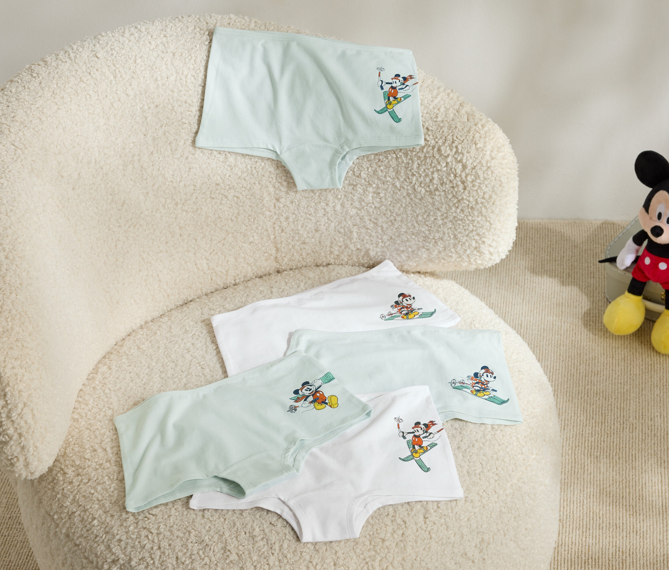Na chlupatém křesle leží čtyři dětské spodky s obrázkem Mickey Mouse na lyžích a vedle křesla sedí plyšový Mickey Mouse.