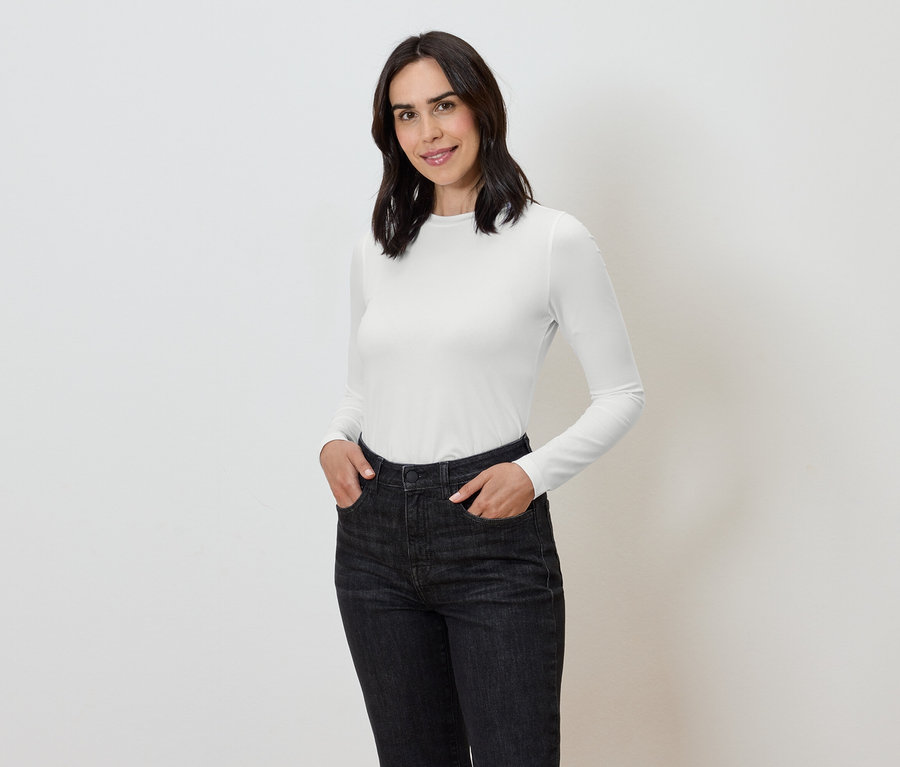 Žena s rukama v kapsách tmavých džínů a bílém tričku s dlouhým rukávem.