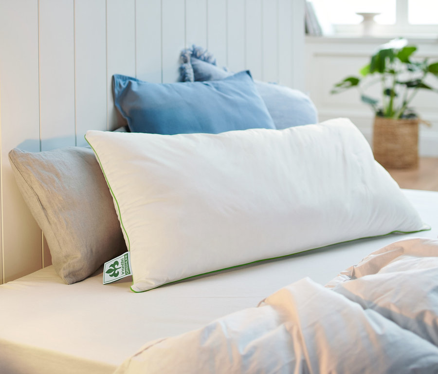 Na posteli leží bílý polštář irisette® greenline o rozměrech cca 80 x 40 cm.