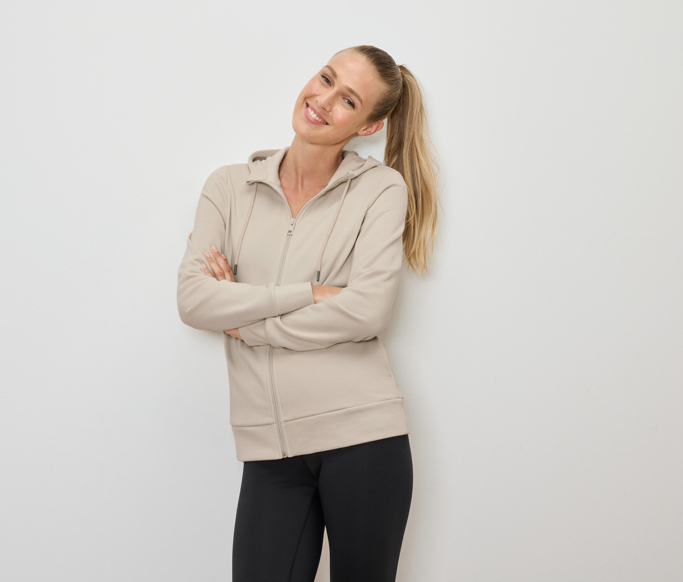 Usmívající se žena s blond vlasy v culíku pózuje s rukama zkříženýma na hrudi, oblečená v béžové mikině na zip a černých legínách.