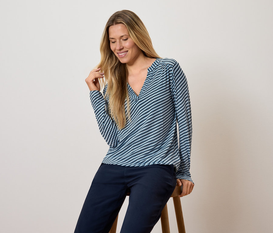 Žena sedí na stoličce, má na sobě modrobílý pruhovaný top s dlouhým rukávem a tmavě modré kalhoty.