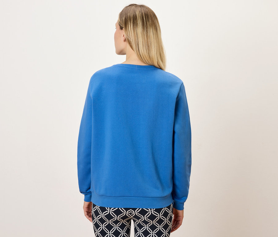 Zezadu je vidět žena s blond vlasy v modré mikině a vzorovaných kalhotách.