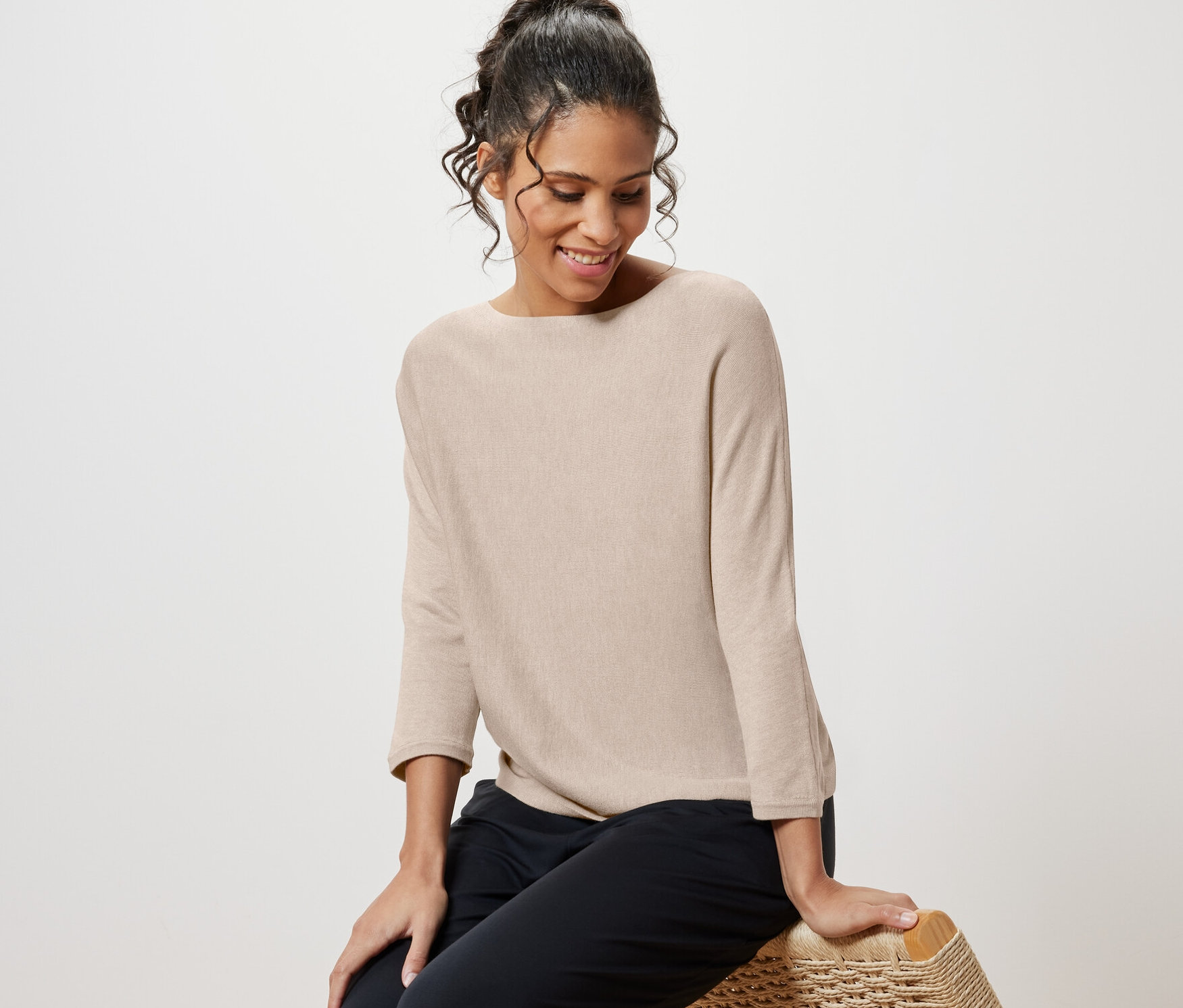 Žena sedí na proutěném křesle a má na sobě béžový melírovaný svetr z jemného úpletu.