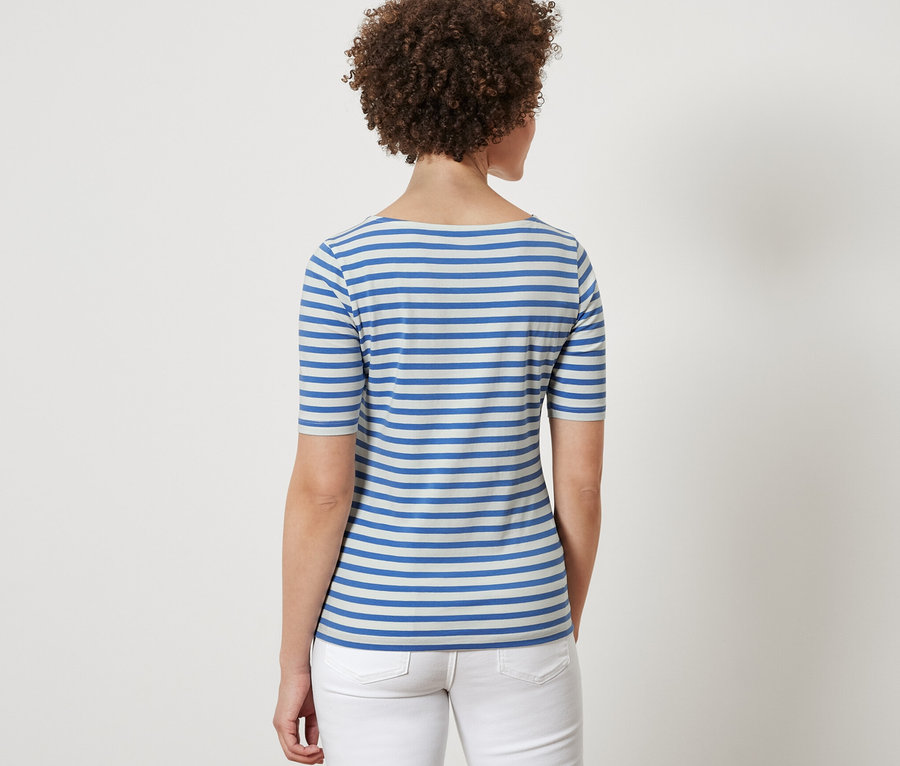 Zezadu je vidět žena s hnědými kudrnatými vlasy, která má na sobě modrobílé proužkované triko s polodlouhým rukávem a bílé džíny.