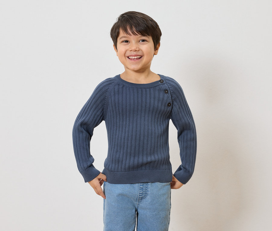 Usmívající se chlapec s hnědými vlasy má na sobě tmavě modrý žebrovaný svetr s knoflíky a světle modré džíny.