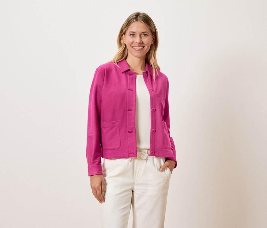 Žena má na sobě růžovou bundu přes bílý top a krémové kalhoty.