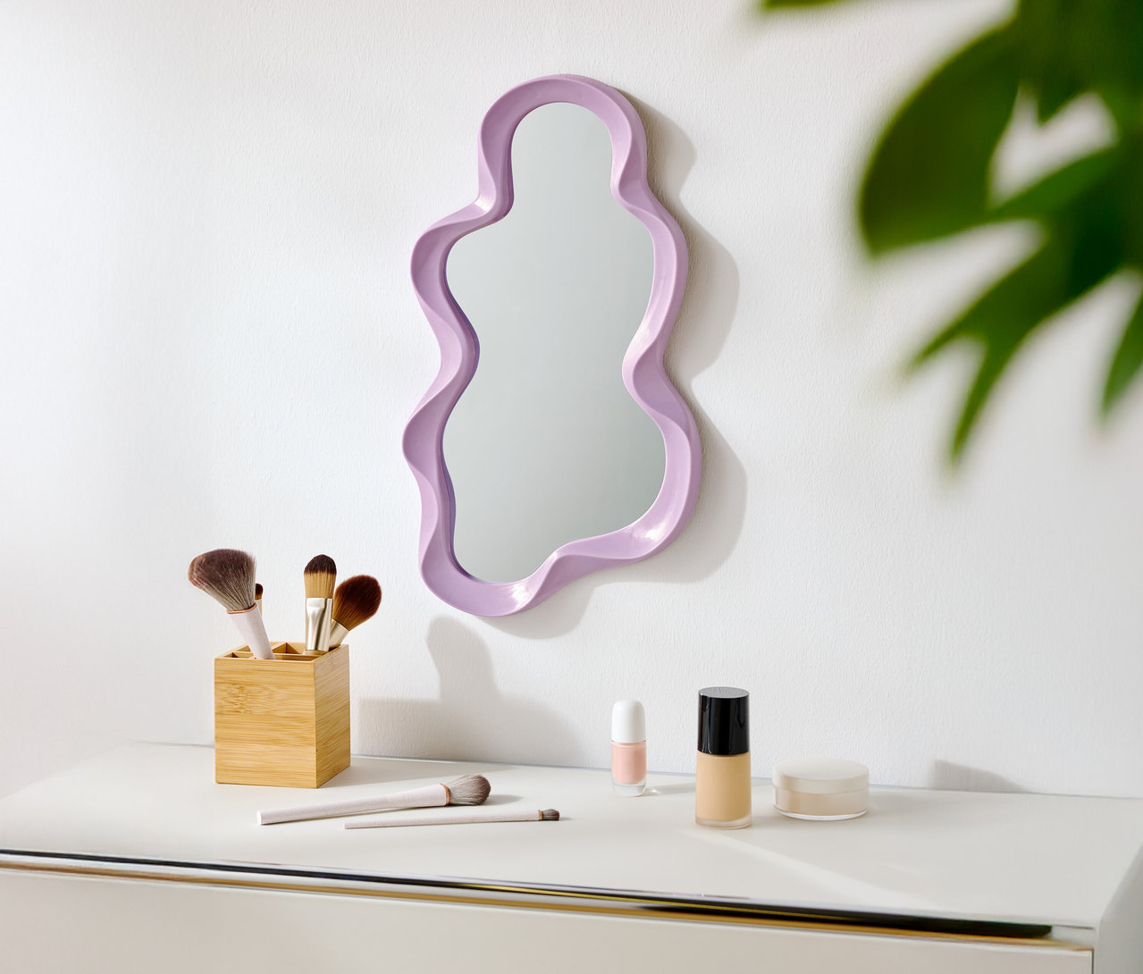 Na bílé stěně visí fialové zrcadlo nepravidelného tvaru. Pod ním jsou na bílém stole štětce na make-up a kosmetika.