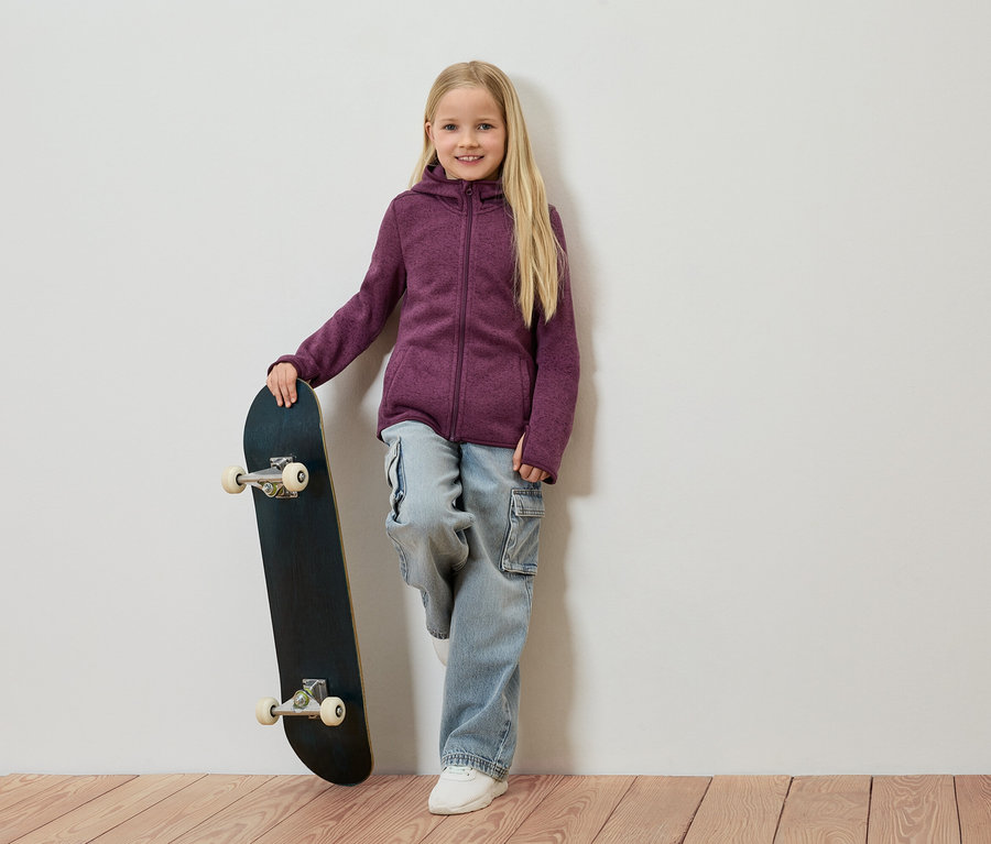 Dívka s blond vlasy se opírá o zeď, drží skateboard a má na sobě fialovou fleecovou bundu, džíny a bílé boty.
