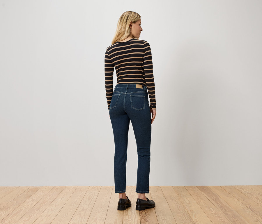 Žena s blond vlasy stojí zády ke kameře a má na sobě pruhované tričko s dlouhým rukávem, modré džíny a černé boty.