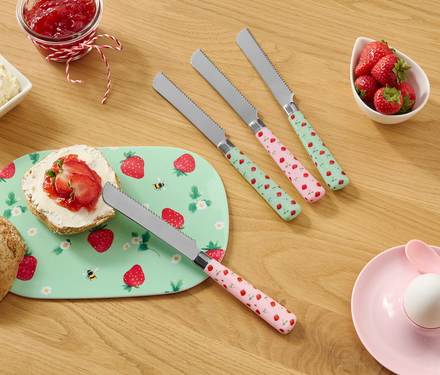 Na dřevěném stole je houska, talíř se vzorem jahod, smetanový sýr, džem, jahody a čtyři nože se vzorem jahod.