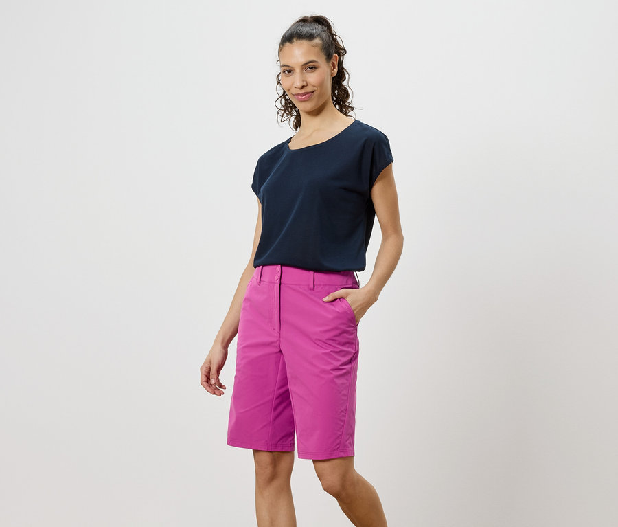 Žena v tmavém tričku a růžových šortkách stojí na bílém pozadí.