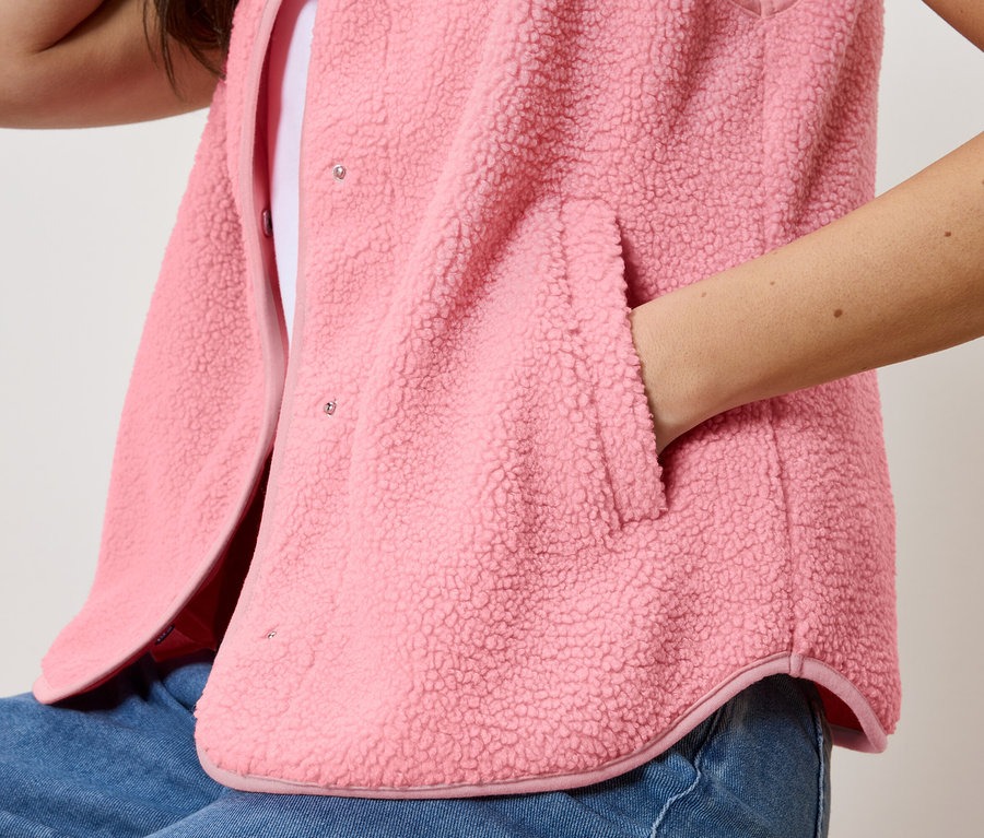 Žena s rukou v kapse plyšové vesty přes bílý top a džíny.